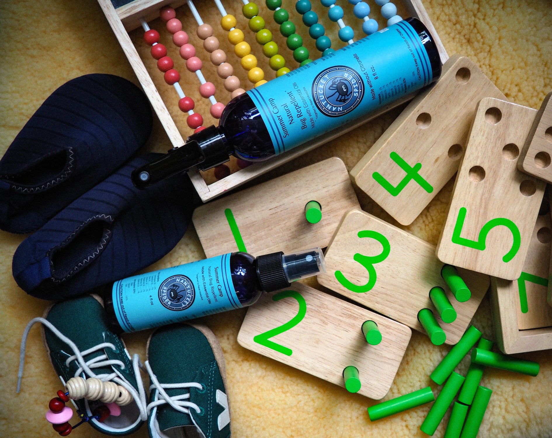 Two blue spray bottles with circular labels are arranged on wooden number boards with green pegs, alongside a pair of dark blue slippers, green sneakers, colorful wooden beads, and green pegs on a textured beige surface.